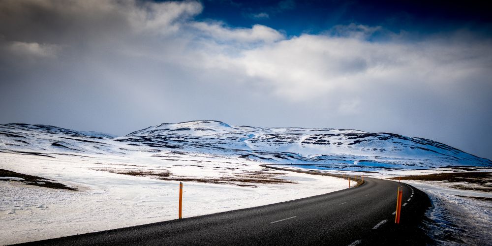 Landscape photograph taken on the Hringivegur ring road in Iceland. This was on the section between Egilsstadir and Akureyri. The road is a ribbon of black tarmac sweeping through a snowy landscape with a rocky hill in the background and a wall of white cloud.