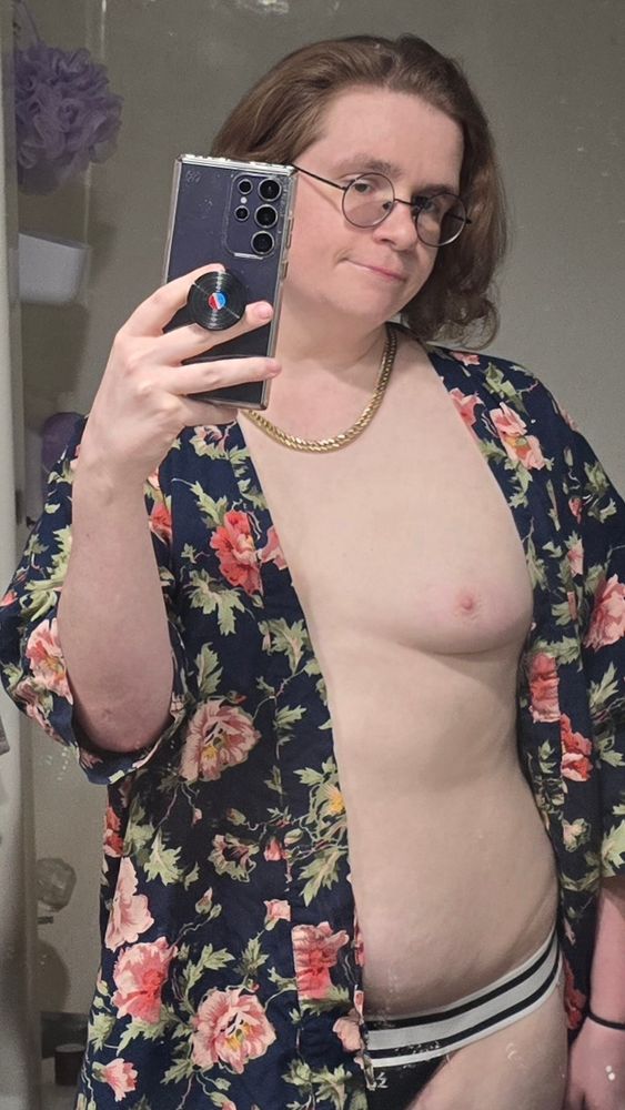 A white nonbinary trans woman takes a mirror selfie, they're have a brown hair that curls around her shoulders, she wears round glasses, a gold chain, a navy floral kimono style top, one of their boobs are showing, they also have a black and white jock strap.
