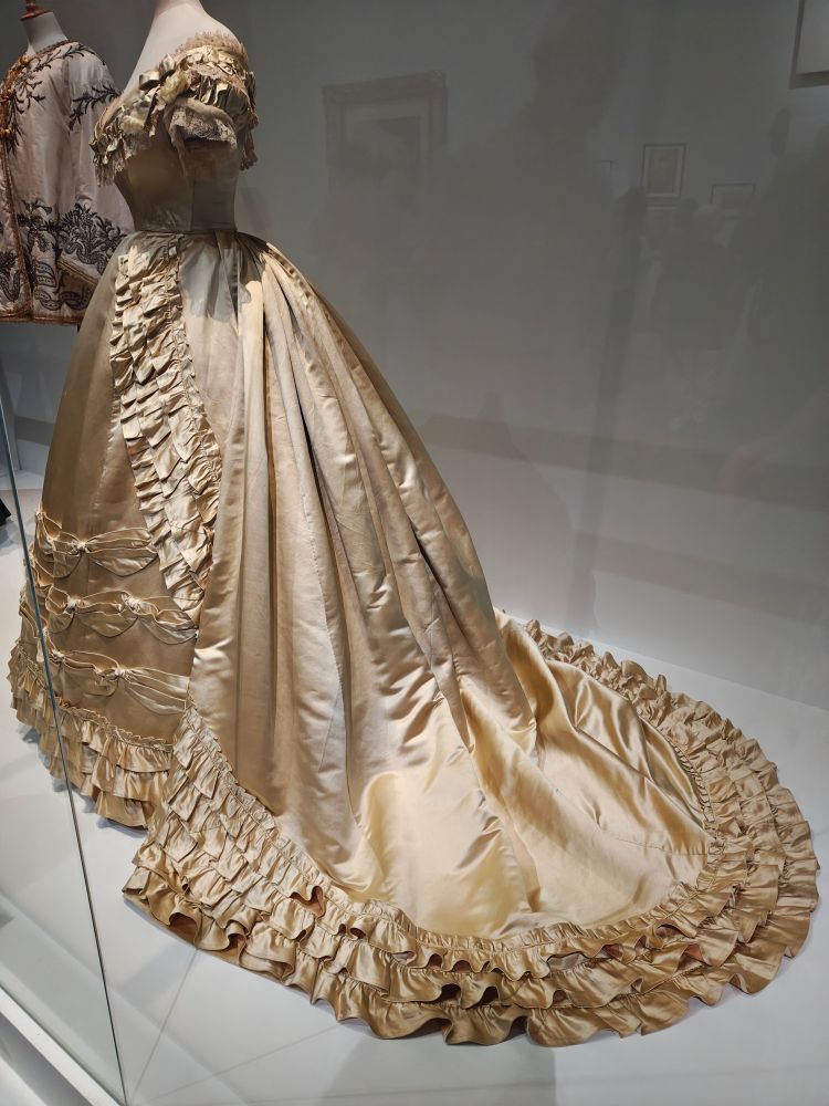 A gold silk-satin late-1860's House of Worth and Boberg gown, with a bustle and train edged in layers of ruffles, with a low-necked evening top. Just delicious fabric details on the front of the skirt, with three layers of little sideways, I don't know, swags? and more ruffles at the bottom.