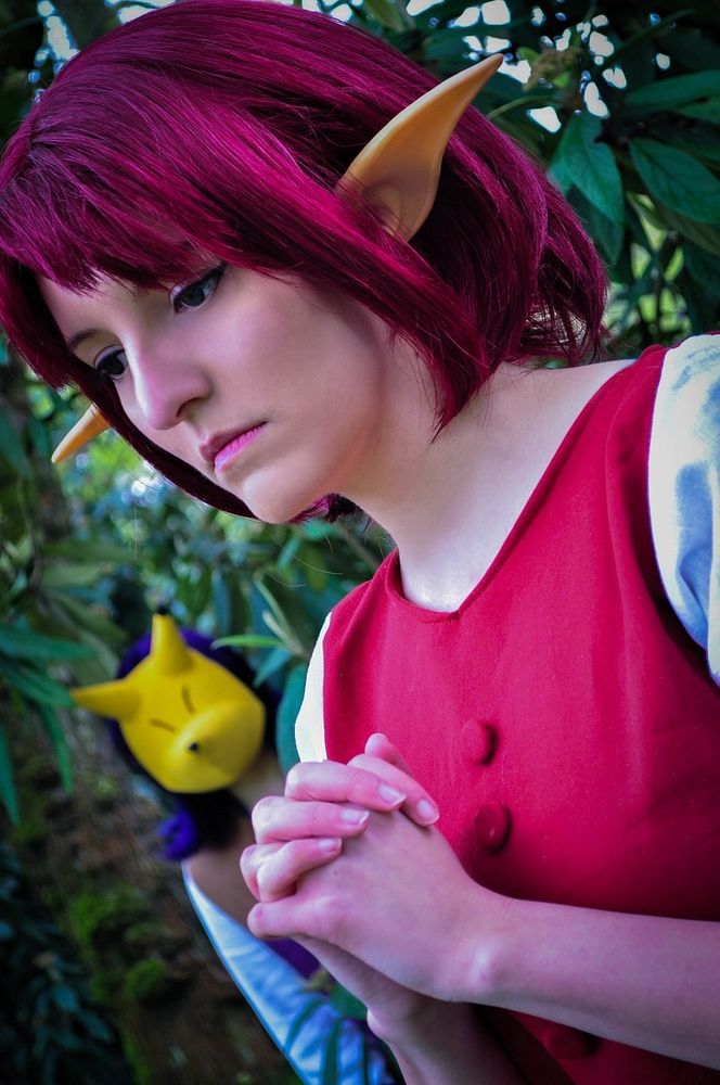 A red haired woman stands there in a praying pose. In the bushes of the background there's a boy hiding from her with purple hair wearing a fox mask. 