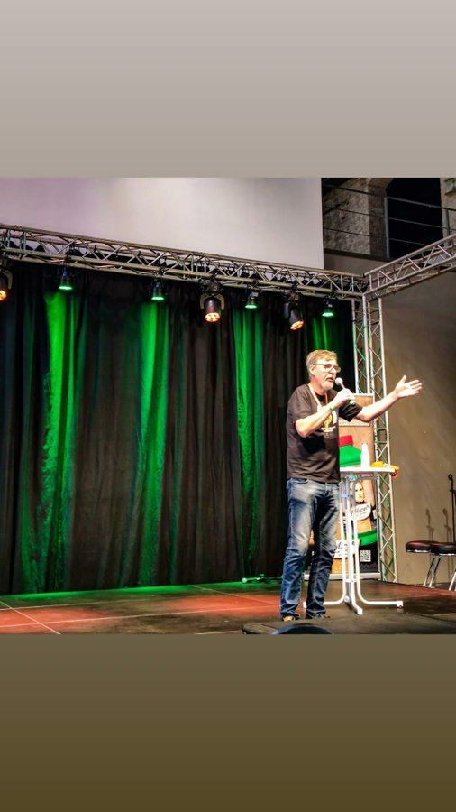 Mann steht auf einer Bühne vor einem grünen Vorhang und hält eine Rede.