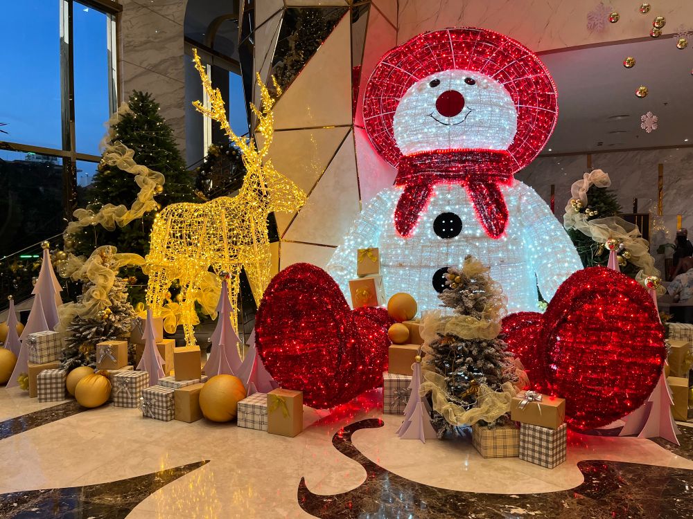 Eine beleuchtete Weihnachtsdekoration bestehend aus Rentier und Schneemann. Davor und daneben goldfarbene Tannenbäume, Kugeln und Geschenke. 