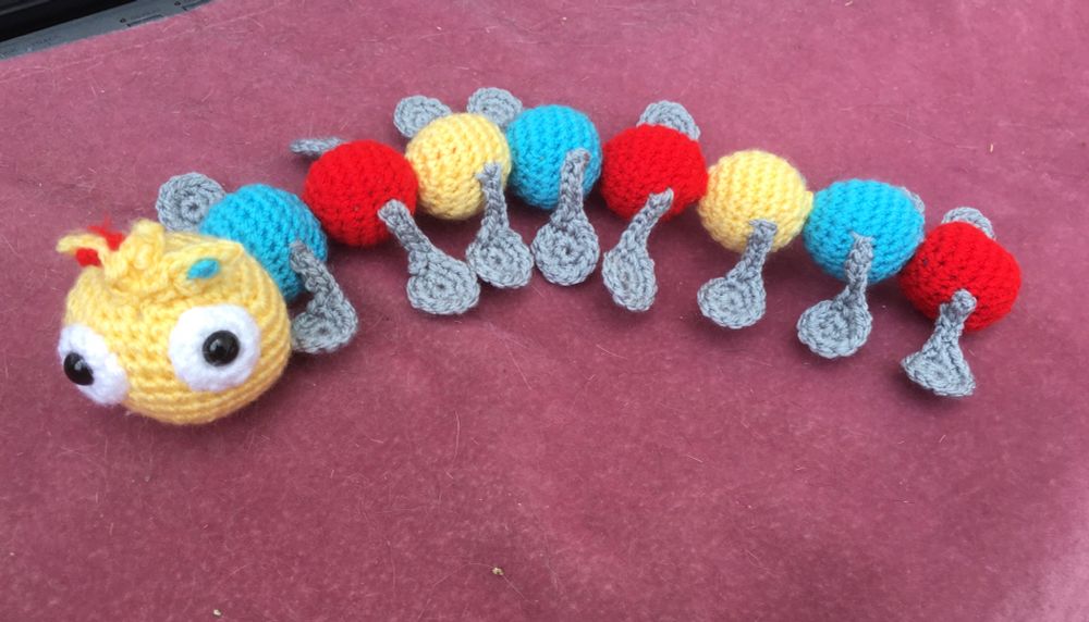 Centipede made of coloured crochet balls in yellow, blue and red. The largest one is the head, it has eyes and a crown. All the legs are grey crochet spoons.
