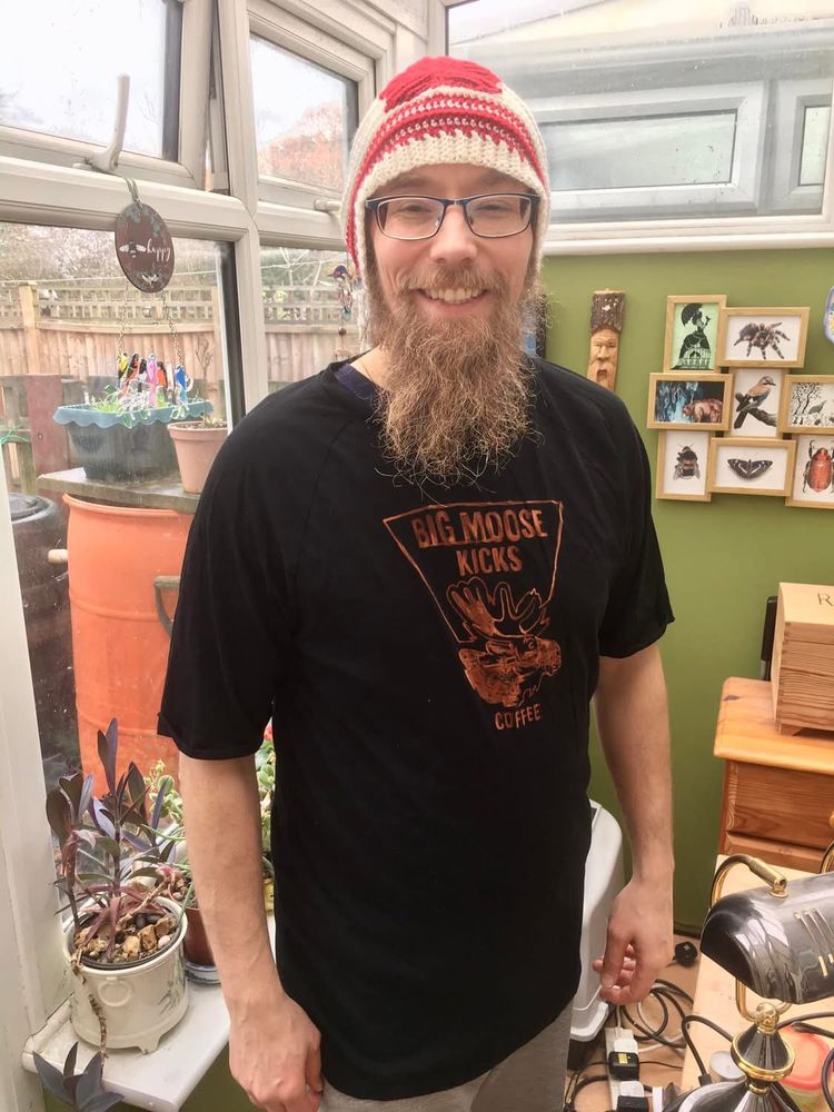 Bearded man in black g shirt with orange design showing a moose and the words Big Moose Kicks Coffee. He also wears a red and cream crochet hat with red maple leaf.