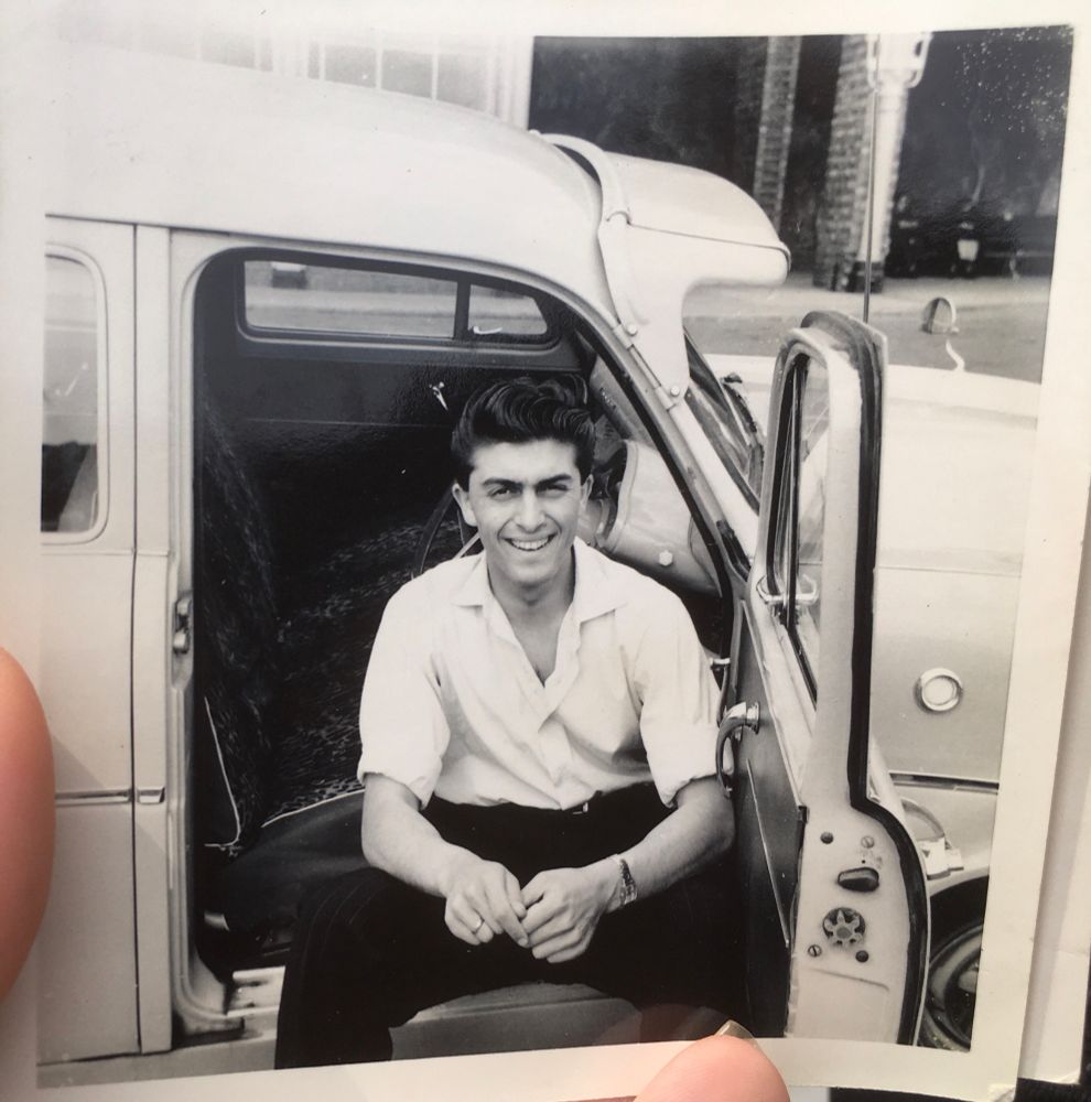 Black and white photo showing a young man with dark hair sitting inside a car sideways with his legs outside and the door open. He wears a white shirt open at the back and black trousers.
