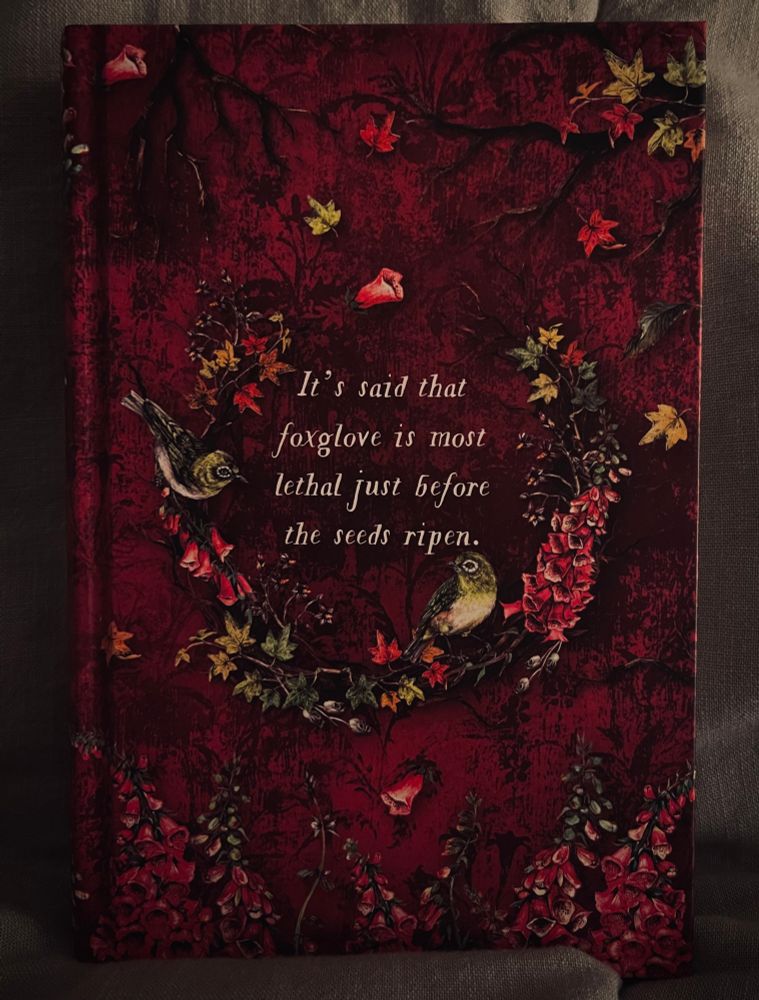 Foxglove book it is red. In the center there is a wreath made of branches wrapped in green leaves and pink and red toned foxglove (bell-shaped flower) there are two little birds perched on the wreath. In the center are the words “It’s said that foxglove is most lethal just before the seeds ripen” 