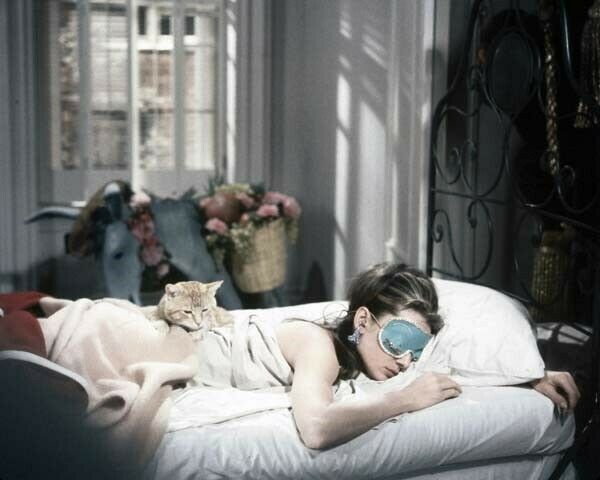 A woman sleeping with a blue eye mask and an orange cat laying on her back