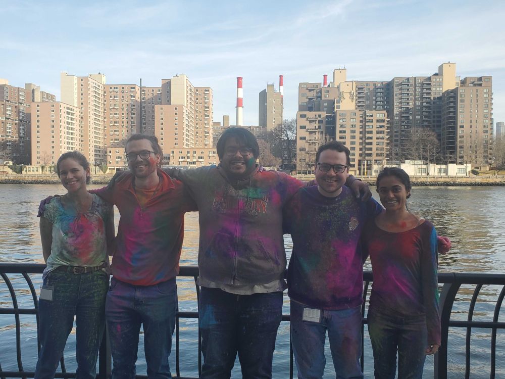 Five people, covered in bright Holi powder, stand arm-in-arm in front of a waterfront railing with a cityscape in the background. They’re all smiling and wearing colorful, powder-stained clothes.
