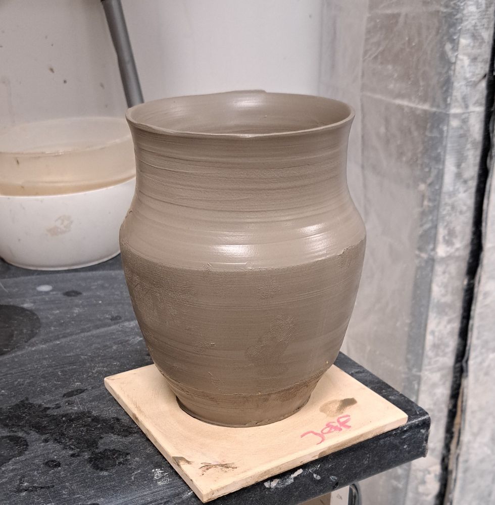A clay vase, straight off the pottery wheel. Pictured in a studio, sitting on a bisque tile so it can dry and firm up.