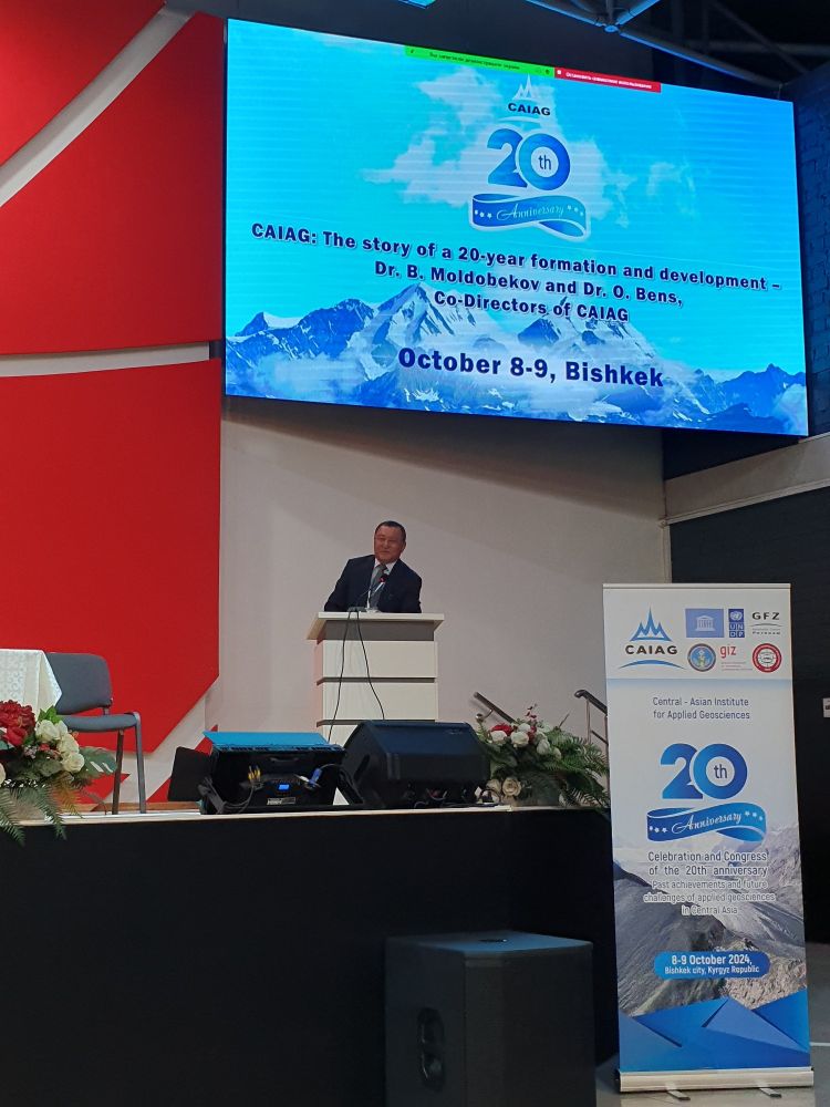 Dr Bolot Moldobekov, the co-director of CAIAG, standing on stage. Behind him is a slide announcing his presentation with co-director Dr Oliver Bens. To the right a banner with 20th anniversary highlighted