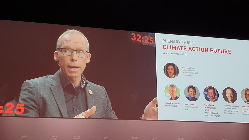 Photo of a wallscreen showing Johan Rickström speaking engaged, with his hands somewhat spread out. To his right are small photographs of the faces of the moderator and the five panellists.