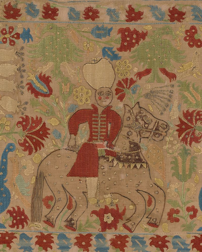 Embroidered cushion cover showing a man riding a horse, surrounded by decorative motifs of flowers.