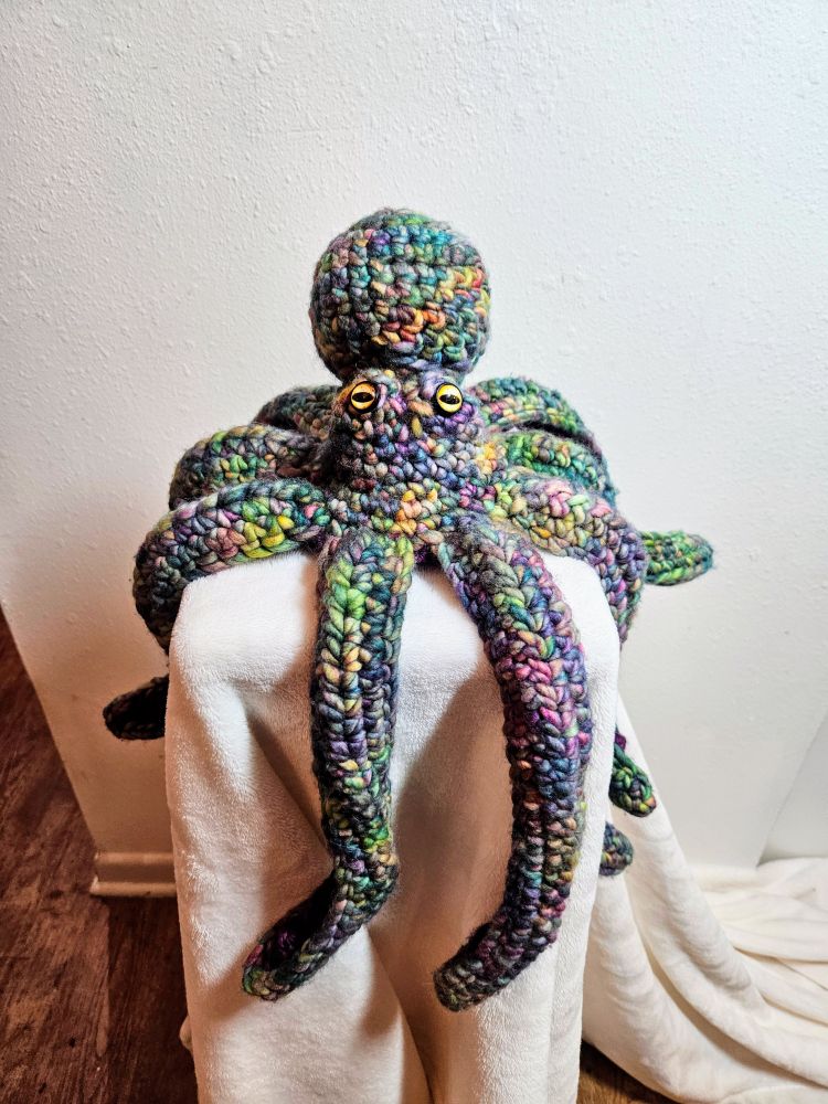A crocheted octopus pictured from the front. She is made of multicolored yarn. Various shades of purple and green are the most common, but there are also spots of yellow and orange. She has yellow eyes with horizontal slit-like black pupils. Her tentacles curl, and she has a giant, bulbous head.