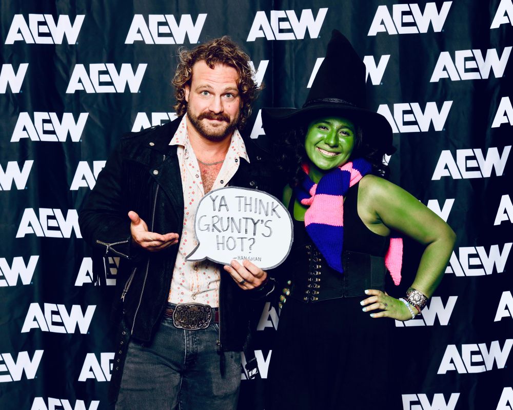 picture of me (emi!) with hangman adam page at starrcast. i am in full cosplay of Gruntilda from Banjo Kazooie the videogame, and hangman is dressed so sexy with his leather jacket and an open white pearlsnap. he is holding a little speech bubble sign that reads, “Ya think Grunty’s hot?” which he said in a stream with Evil Uno while playing Banjo Kazooie