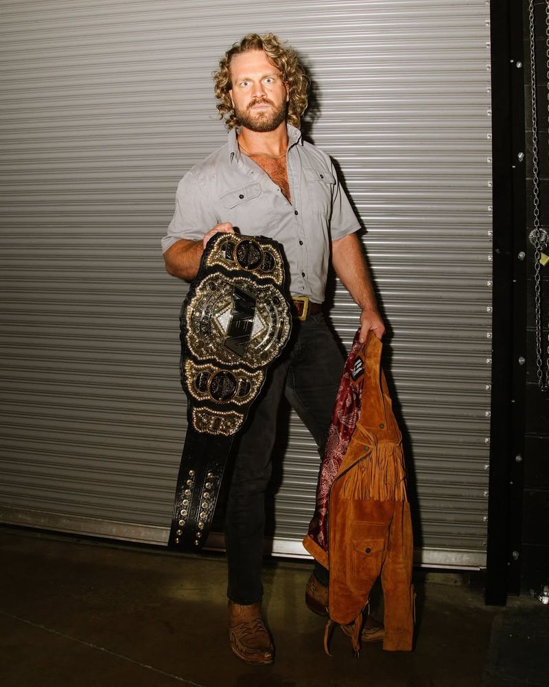 hangman adam page holding his aew men’s world championship with one hand. grey garage door background. he is wearing a grey button down unbuttoned halfway down his chest, brown belt, dark jeans. he is holding his brown suede leather jacket in the other hand. he is looking straight at the camera. all his weight is on his right leg.