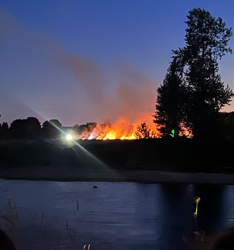 A dark river in front, a riverbank, then a line of tall flames and darkish sky