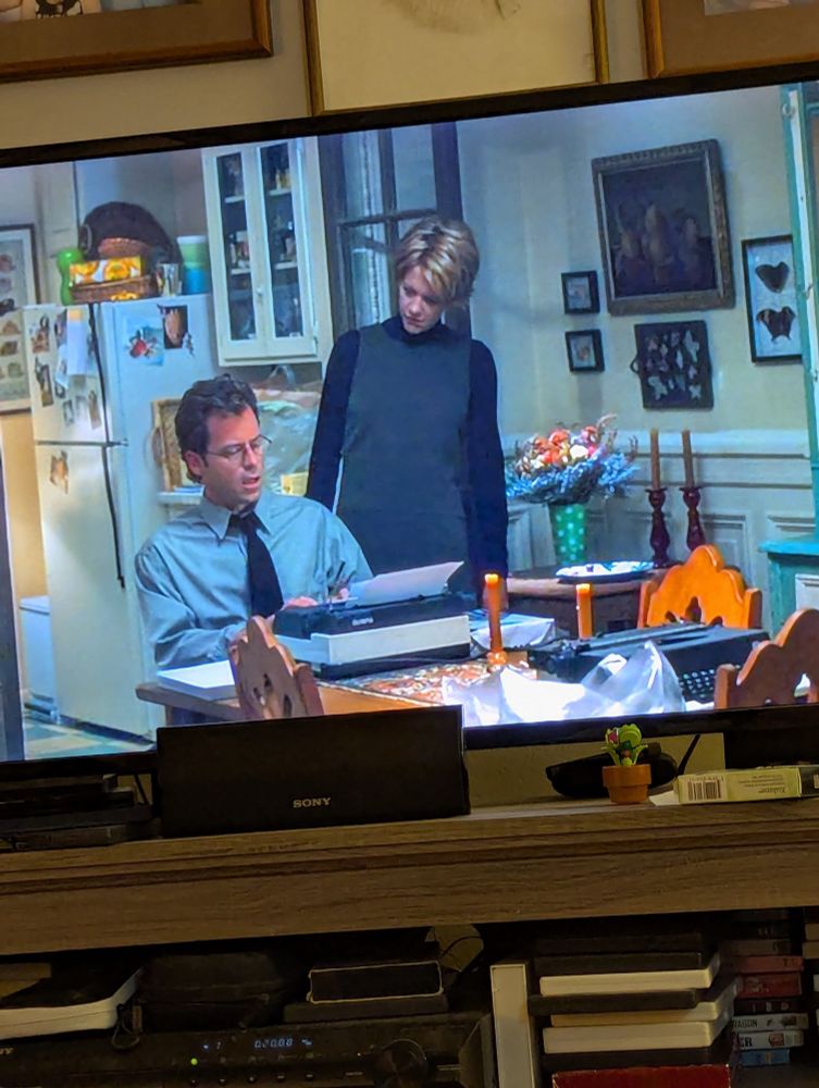 Screenshot of You've Got Mail Meg Ryan looking at Frank type on a typewriter with another typewrite in the frame