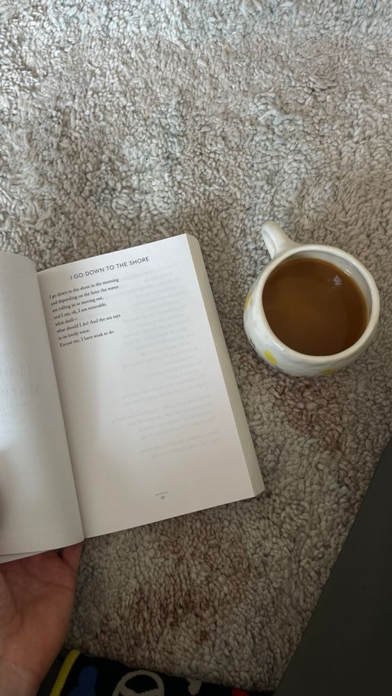 A picture of a cup of coffee and an open book. The book is called Devotions by Mary Oliver and the poem “I Go Down to the Shore”.
