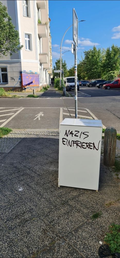 Eine Berliner Straßenecke im Sonnenlicht. Im Vordergrund steht ein Kühlschrank, auf den jemand mit Edding 