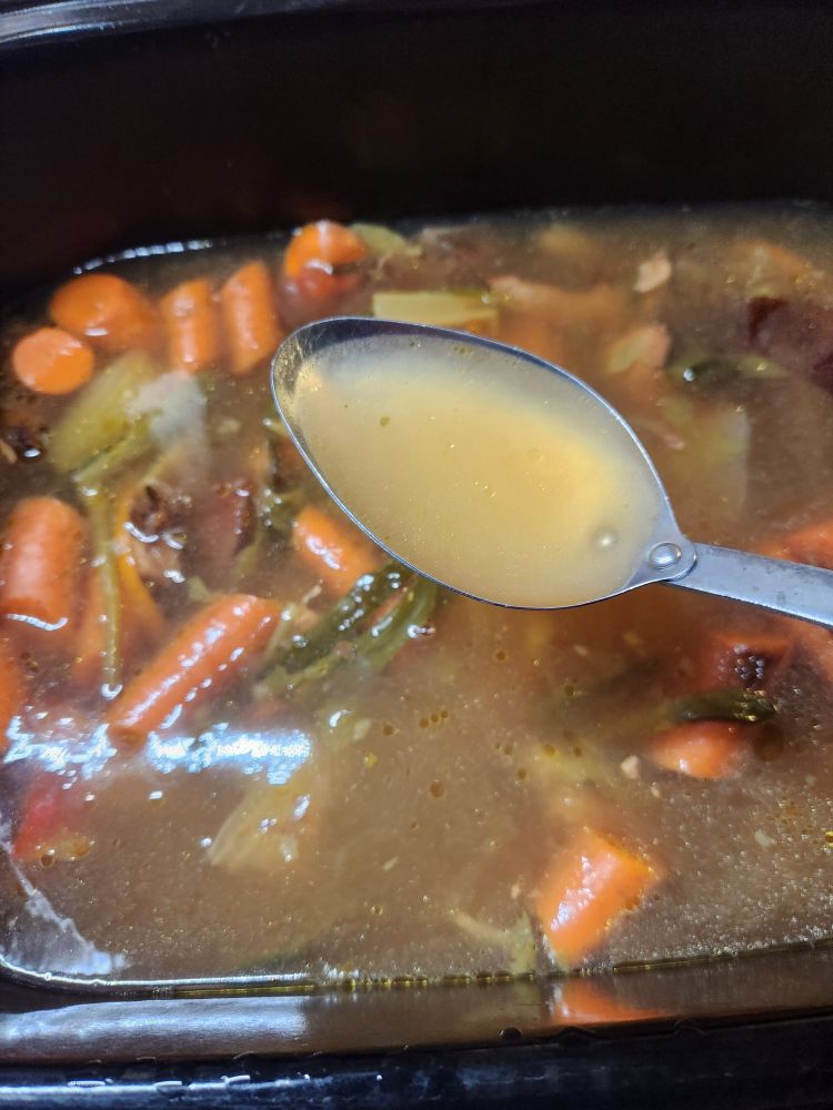 Overhead view of an electric roaster, the roaster is full of stock and vegetables. A spoon is in the center of the image, it's holding a spoonful of clean broth