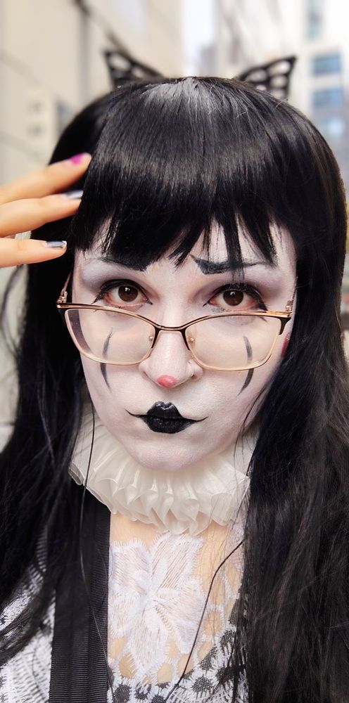 Selfie Photo of a tall woman dressed as a demure and regal mime, looking pensive and sweet at the camera. She's got black streaks on her cheeks, a red button nose, and thin glasses w a butterfly hair clip on the back of her long black hair