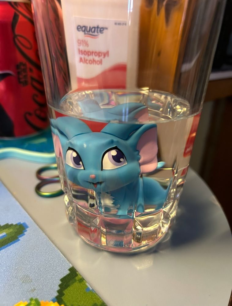 A small figure of a blue acara neopet submerged in a glass of clear liquid, a bottle labeled “91% isopropyl alcohol” is visible behind.
