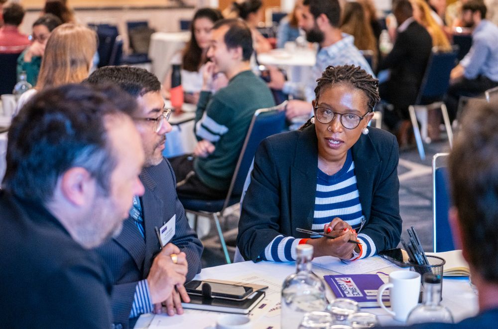 Conference or meeting setting with multiple groups of people seated around tables, engaged in conversation. Tables contain notebooks, pens, cups, and water bottles, suggesting an organised group activity or discussion.