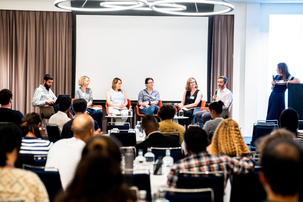 Panel discussion at a conference with six seated speakers on stage and one person standing at a podium. Audience members are seated and facing the stage.