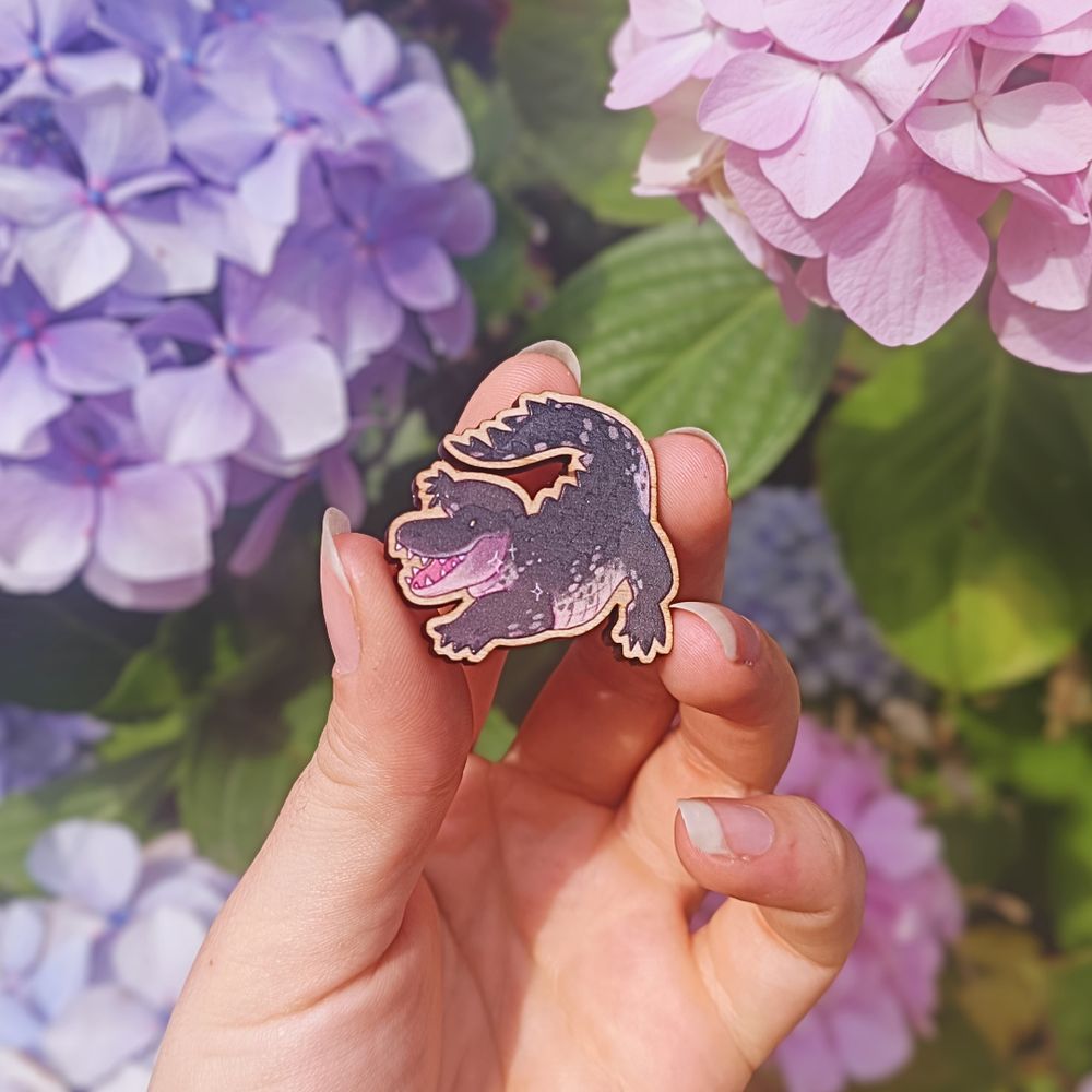 a wooden pin badge of a chibi alligator, held in front of some pink, blue, and purple hydrangeas