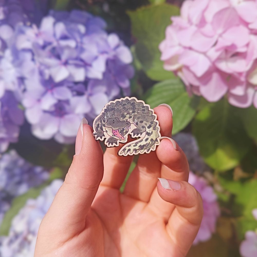 a wooden pin badge of a chibi crocodile, held in front of some pink, blue, and purple hydrangeas