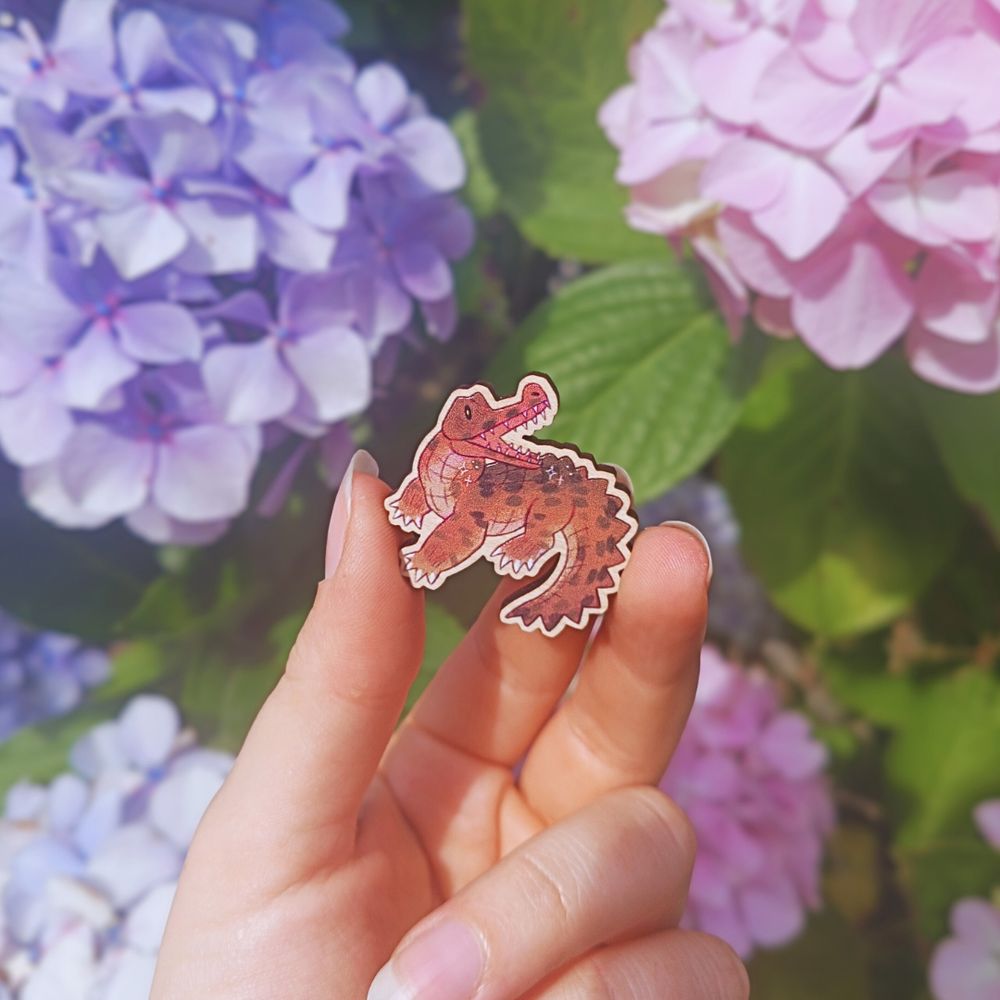 a wooden pin badge of a chibi gharial, held in front of some pink, blue, and purple hydrangeas