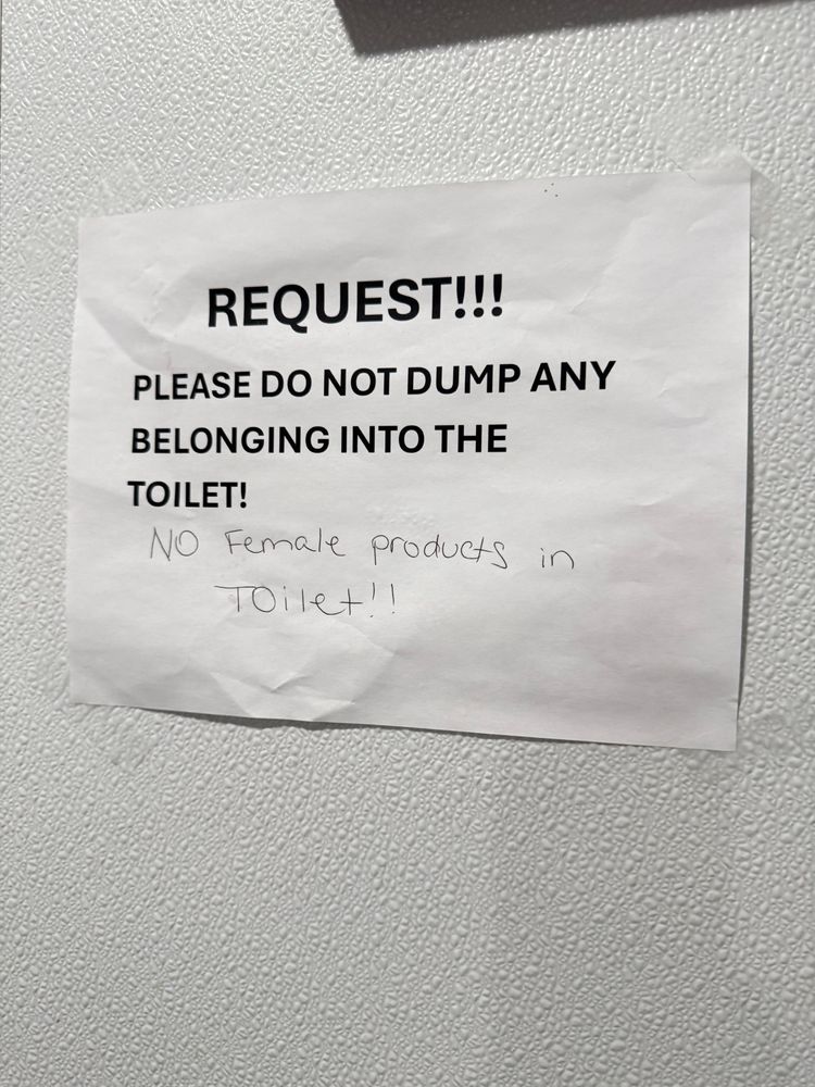 A printed paper sign taped on the wall of a public restroom says: “REQUEST!!! PLEASE DO NOT DUMP ANY BELONGING INTO THE TOILET!” under this handwritten in pen says: “NO female products in toilet!!”