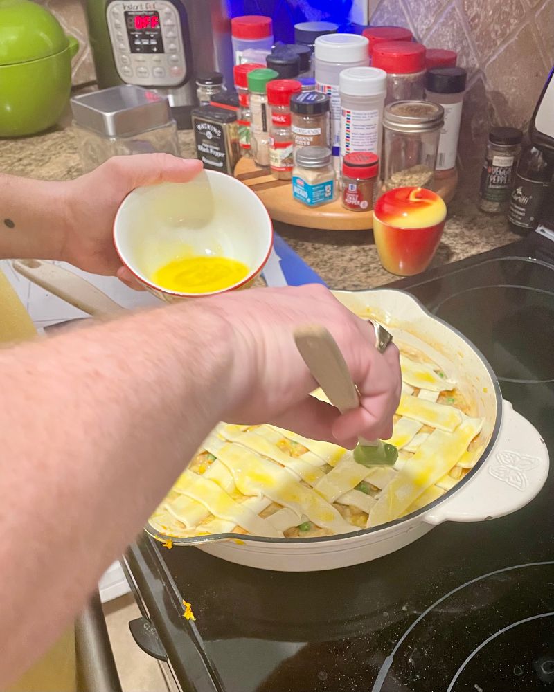 Two disembodied arms brush an egg wash on chickpea pot pie in a white enamel pan. It sits on a black stove, and there are many spices behind it. 