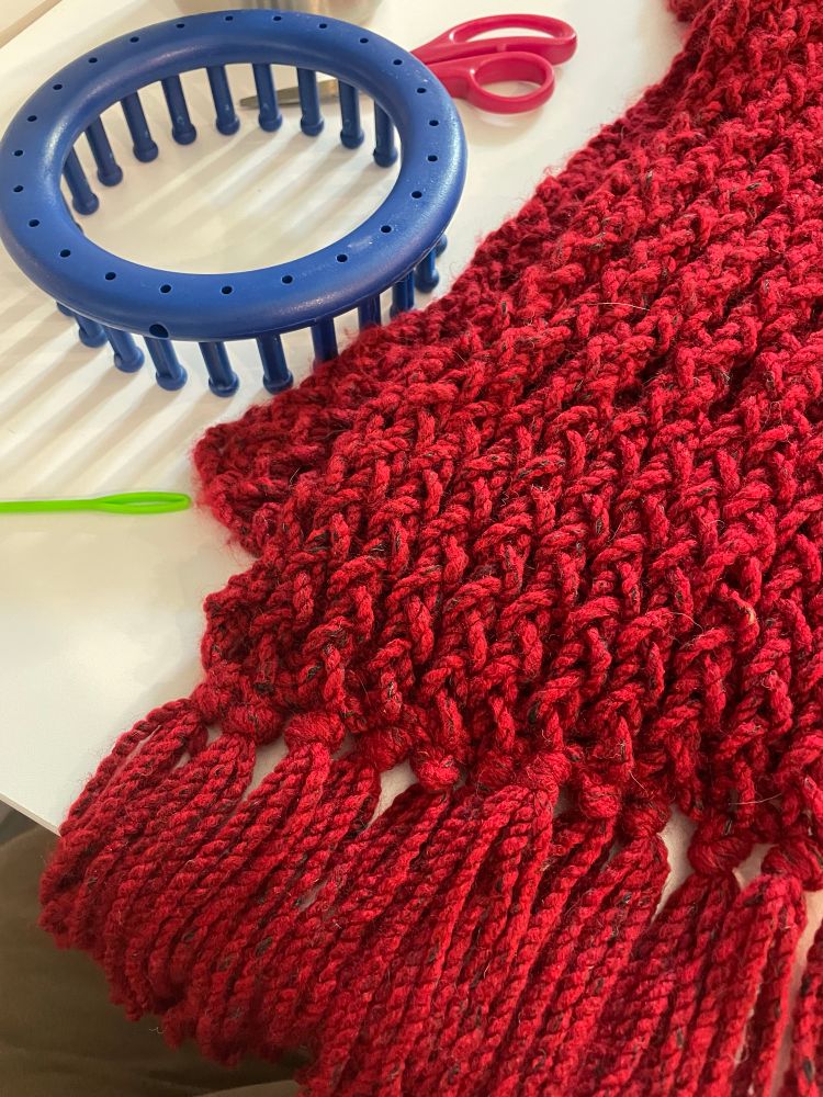 A red scarf with fringe, a blue loom I’m using to measure the fringe, and a neon green needle. 