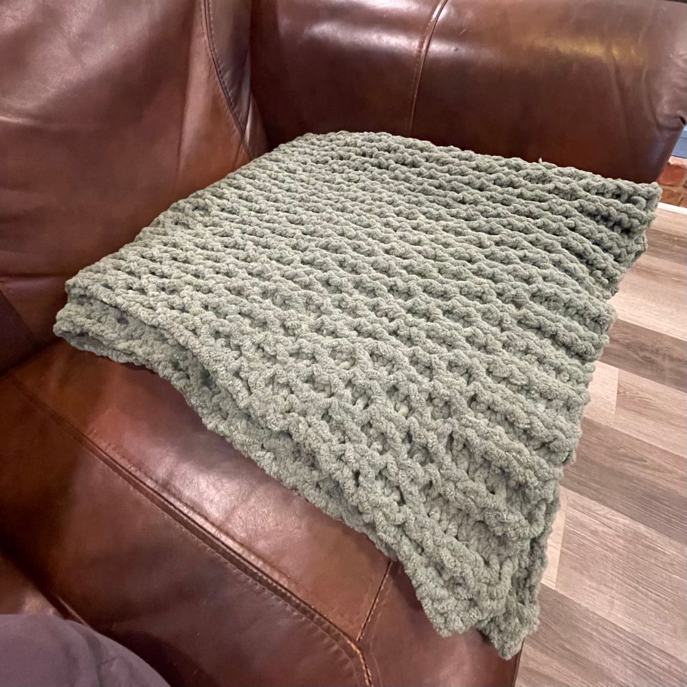A sage green shawl, folded up, with a honeycomb knit. On a brown leather couch. 
