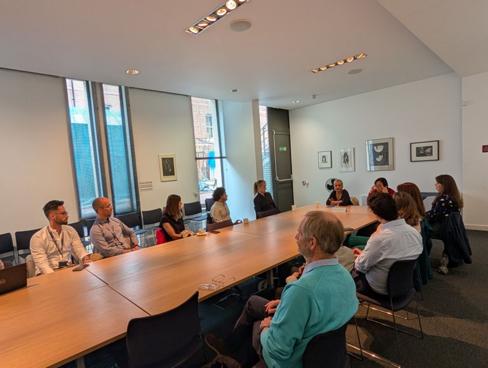 Professor Kamila Hawthorne sits at the head of a large meeting table, engaged in discussion with a group of early and mid-career researchers and DPhil students in a bright, modern seminar room.