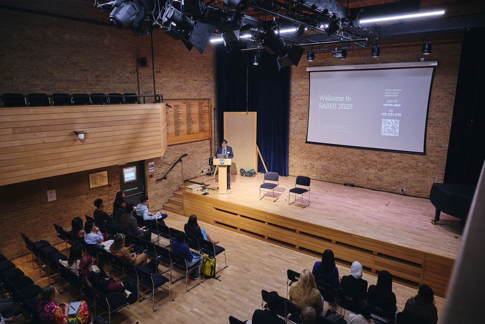 Professor Sir Aziz Sheikh at a podium addressing an audience in a lecture hall, with a welcome slide projected on screen.