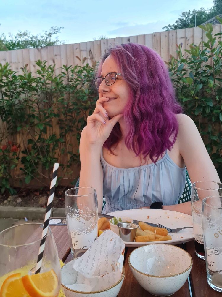 Alfira IRL, sitting at a table in a blue sundress, her hair is purple and wavy. She's looking into the distance with a smile on her face