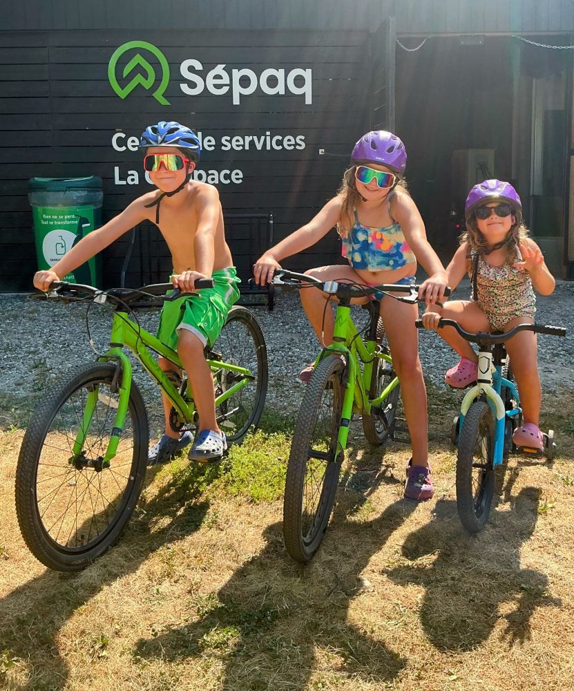 Three young kids pose on bicycles, wearing sunglasses, helmets, and swimsuits.