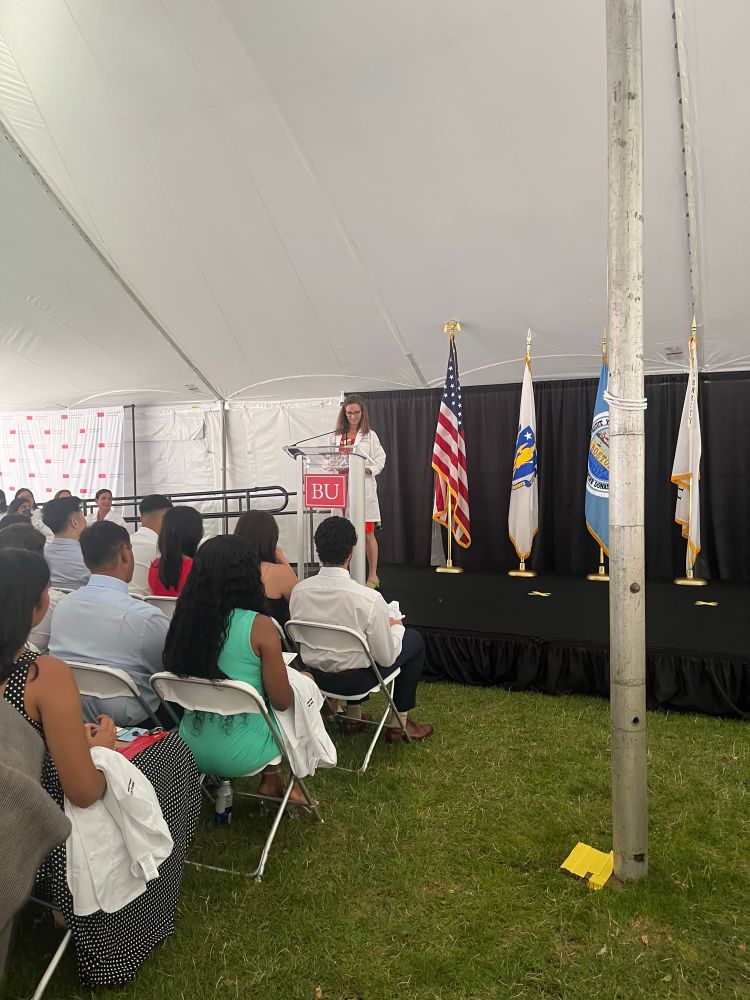 A woman with brown hair and a white coat addresses a crowd of people