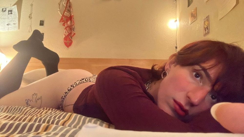 Kit lounging on her bed on her belly in a strong red lip, red sweater dress pulled up to her midriff, cow print thong, and long black socks. Her legs are kicked up and crossed behind her. 