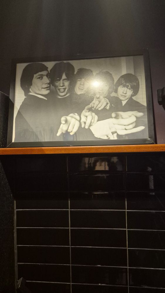 imagem de uma parede de azulejos pretos com uma prateleira e uma foto emoldurada em preto e branco dos rolling stones, eles estão olhando e apontando pra camera e sorrindo
