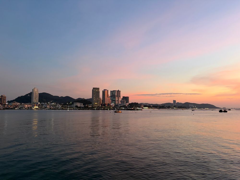 A cluster of high rises in a city by a bay and backed by hills. Boats passing by.