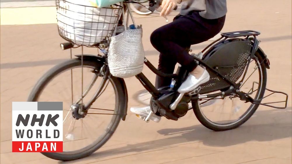 NHK World Japan thumbnail: Person riding an electric-assist step through frame city bicycle equipped with front basket, lights, mudguards, chain guard, rear rack, kickstand. Basket is full of groceries and a handbag hangs from handlebars.
