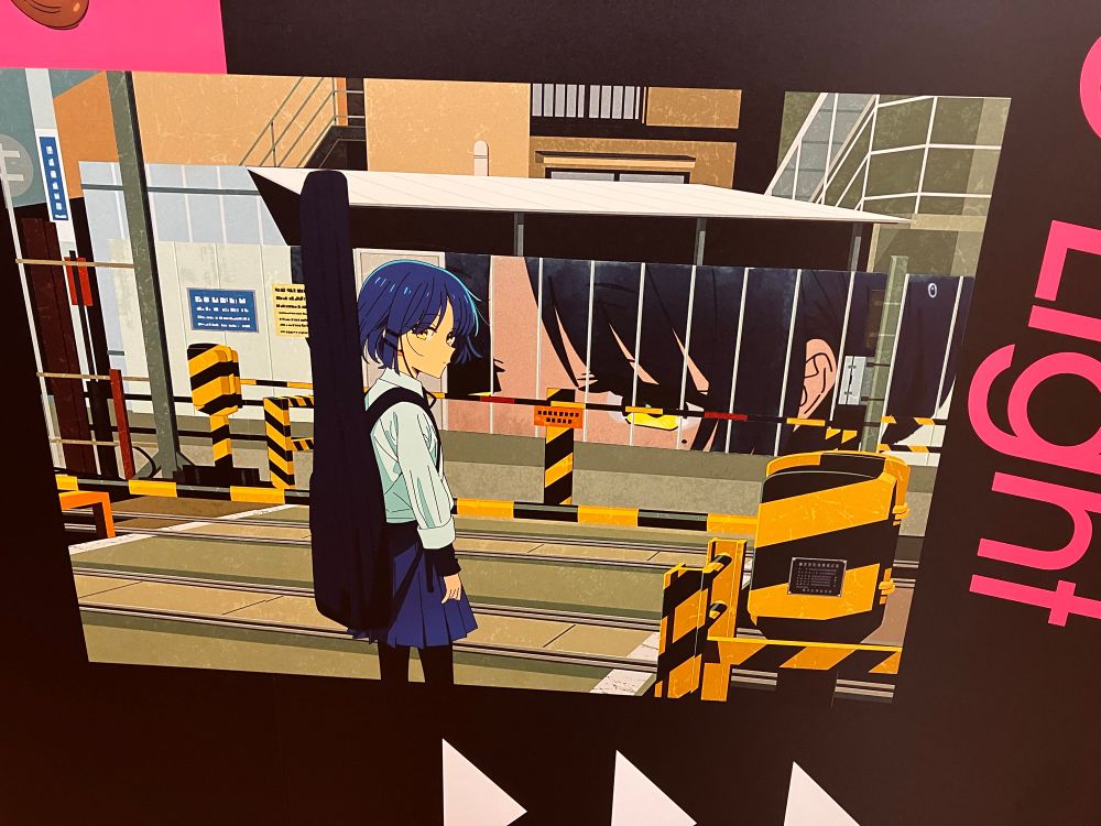 Yamada Ryo in school uniform carrying her bass waits at a railway crossing. A wall on the opposite side features a close up of her eyes.
