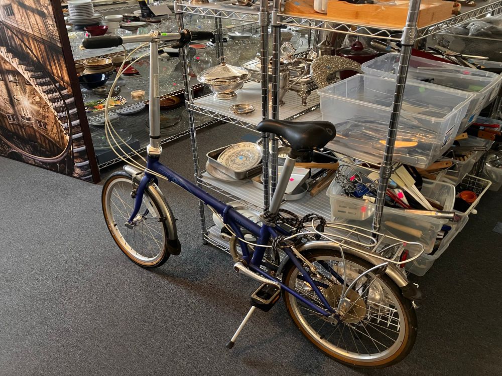 Blue folding bike with mudguards, rack, bungee cords and kickstand in silverware section of store