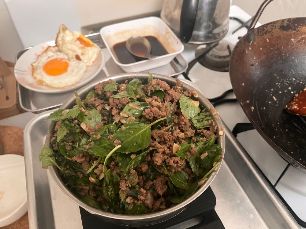 Holy basil beef stir fry in bowl fresh out of wok on gas burner. Fried eggs on plate and sauce on tray over back burner.