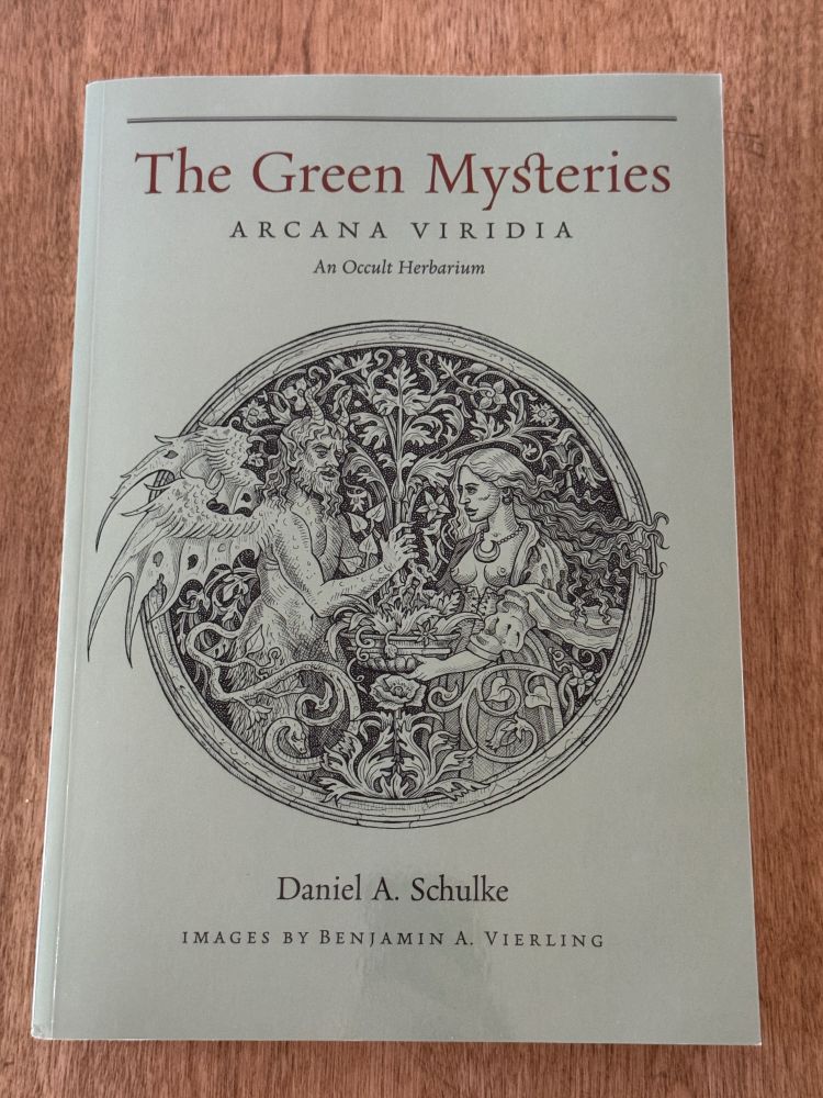 Photograph of a green book on a wooden table. The book has a black and white image of a winged satyr opposite a woman holding a bowl of herbs. The book is titles The Green Mysteries Arcana Viridia An Occult Herbarium by Daniel A. Schulke with images by Benjamin A. Vierling.