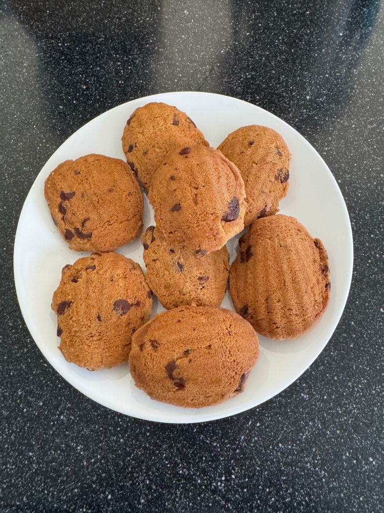 Photograph of a white plate on a black granite background. On the plate are an arraignment of chocolate chip cookies, formed to look like Madelines.