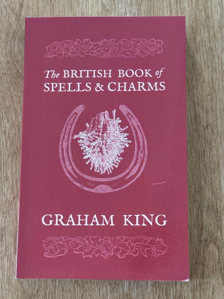 Photograph of a red book with white writing on the cover, with light wood grain visible behind it. The book is titled The British Book of Spells & Charms by Graham King.